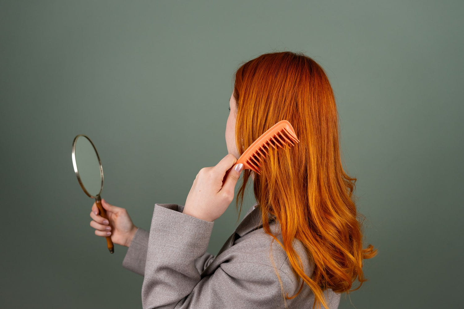 Red-haired woman using a comb while holding a mirror, focusing on hair care and beauty