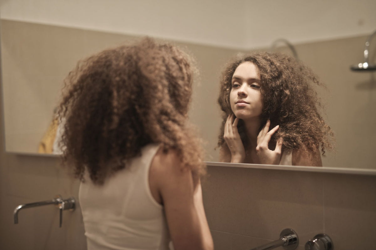 A woman with curly hair contemplates in front of a bathroom mirror, reflecting on her thoughts