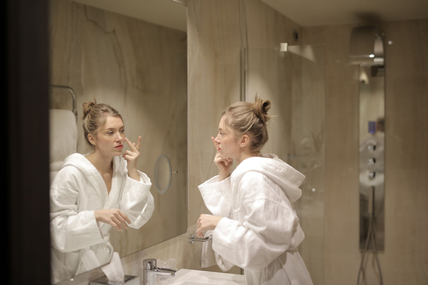 A woman in a white bathrobe applies moisturizer while looking at her reflection in the bathroom mirror
