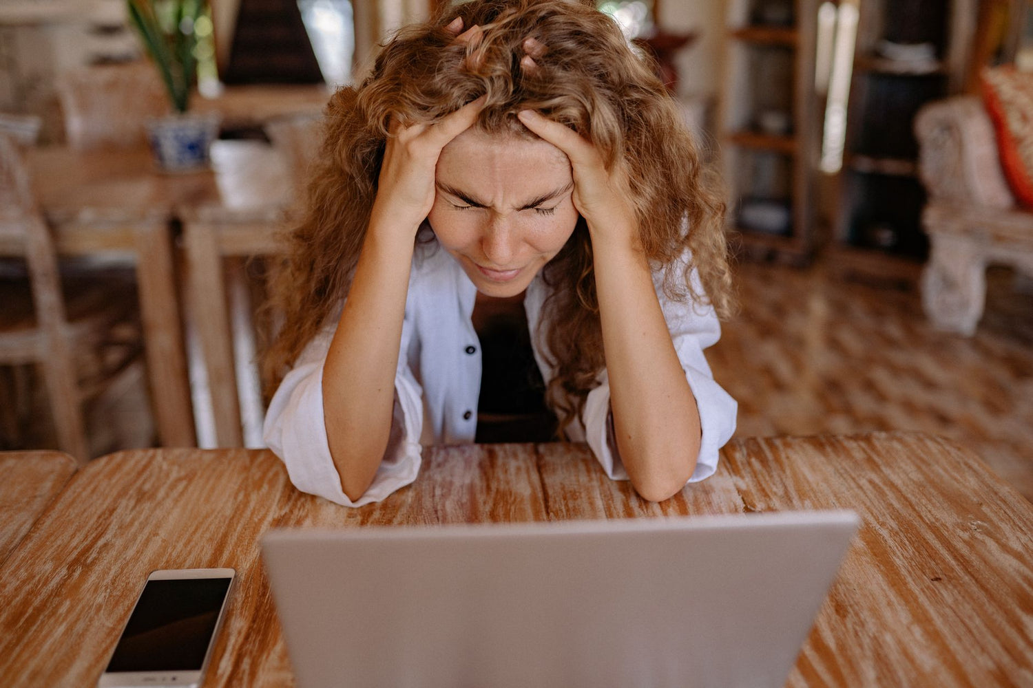 A stressed woman sits at a wooden table, frustrated with her laptop work