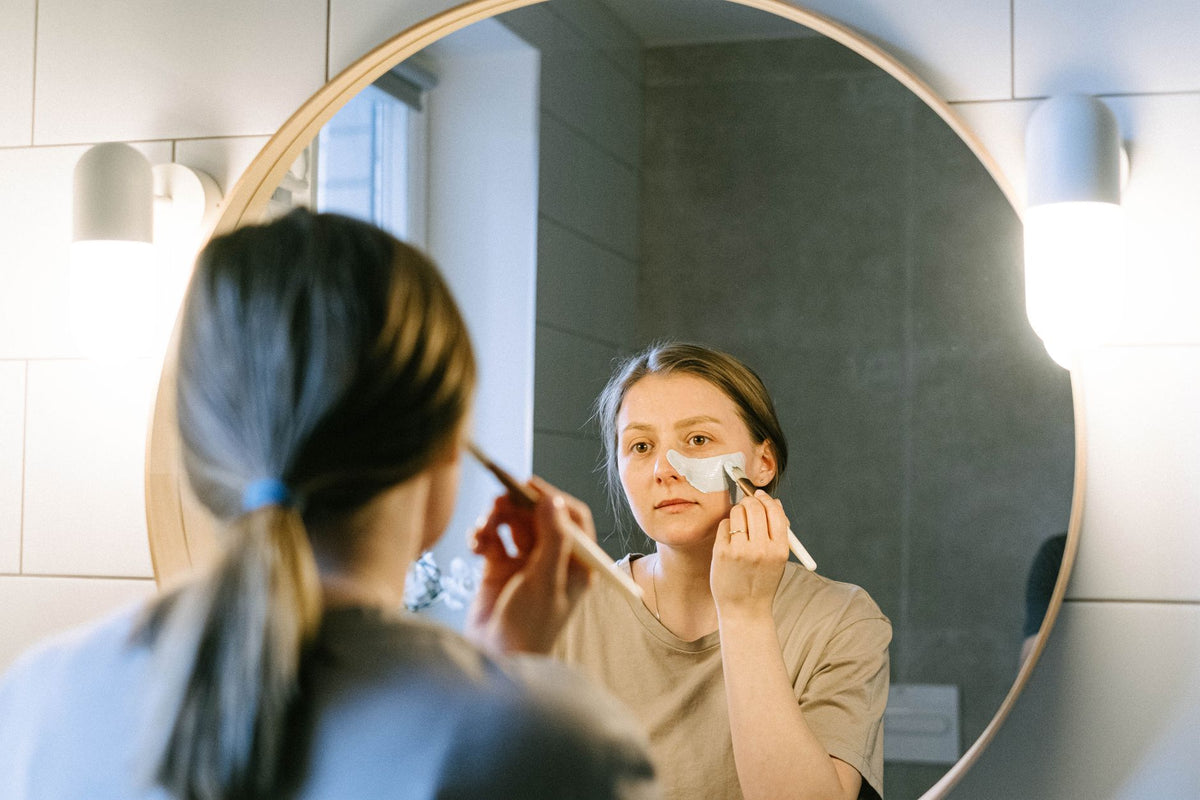 A woman applies facial cream in front of a round bathroom mirror, illustrating a beauty routine
