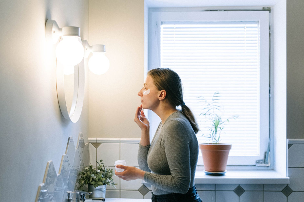 A woman applies face cream in a well-lit bathroom, emphasizing skincare routine