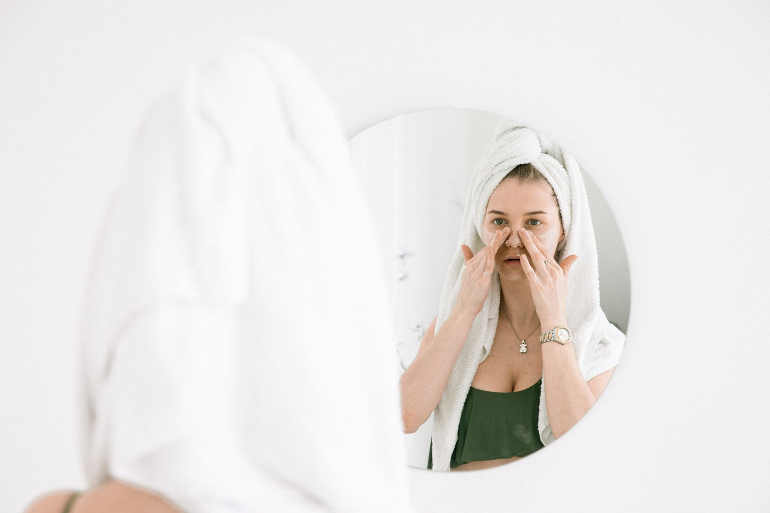 A woman in a towel performing her skincare routine in front of a bathroom mirror