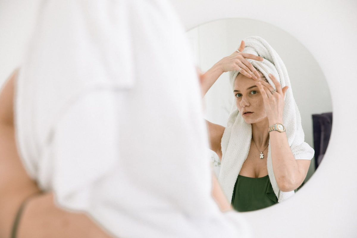 Woman with towel on head applying skincare, enjoying beauty routine in front of mirror