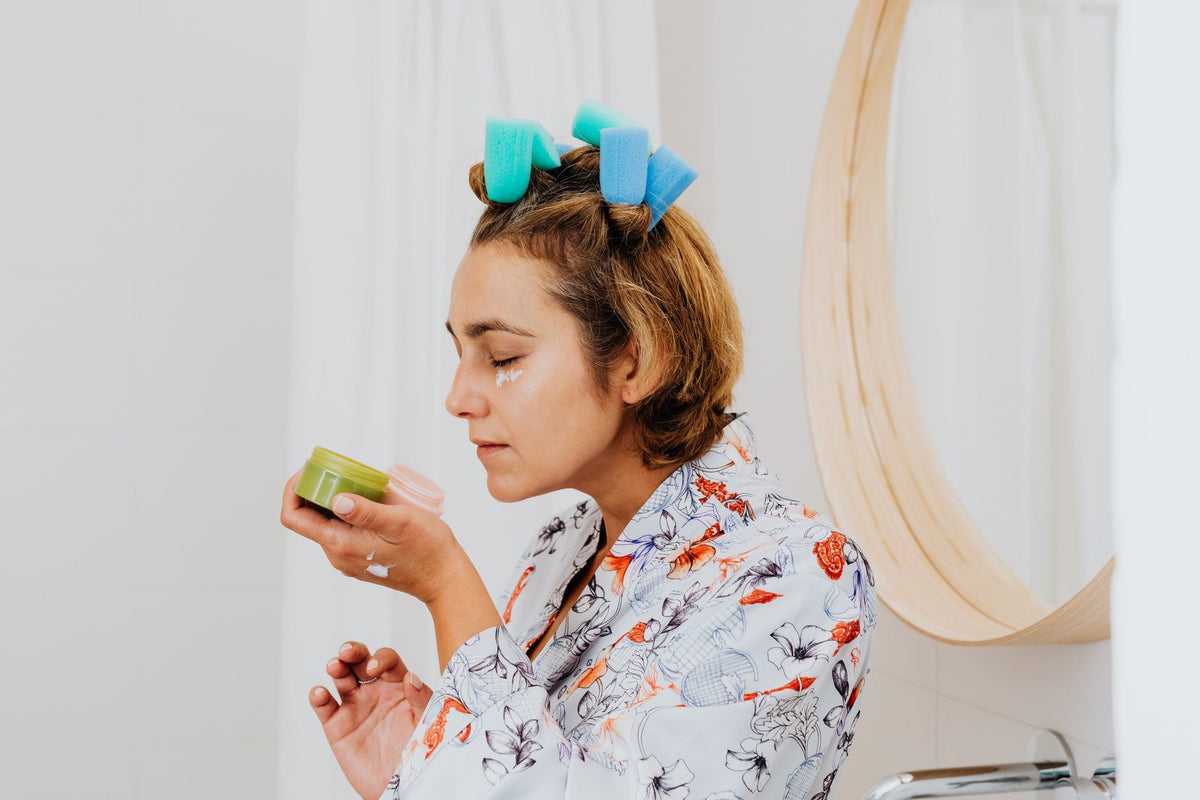 Woman with hair curlers applies skincare cream in front of a bathroom mirror