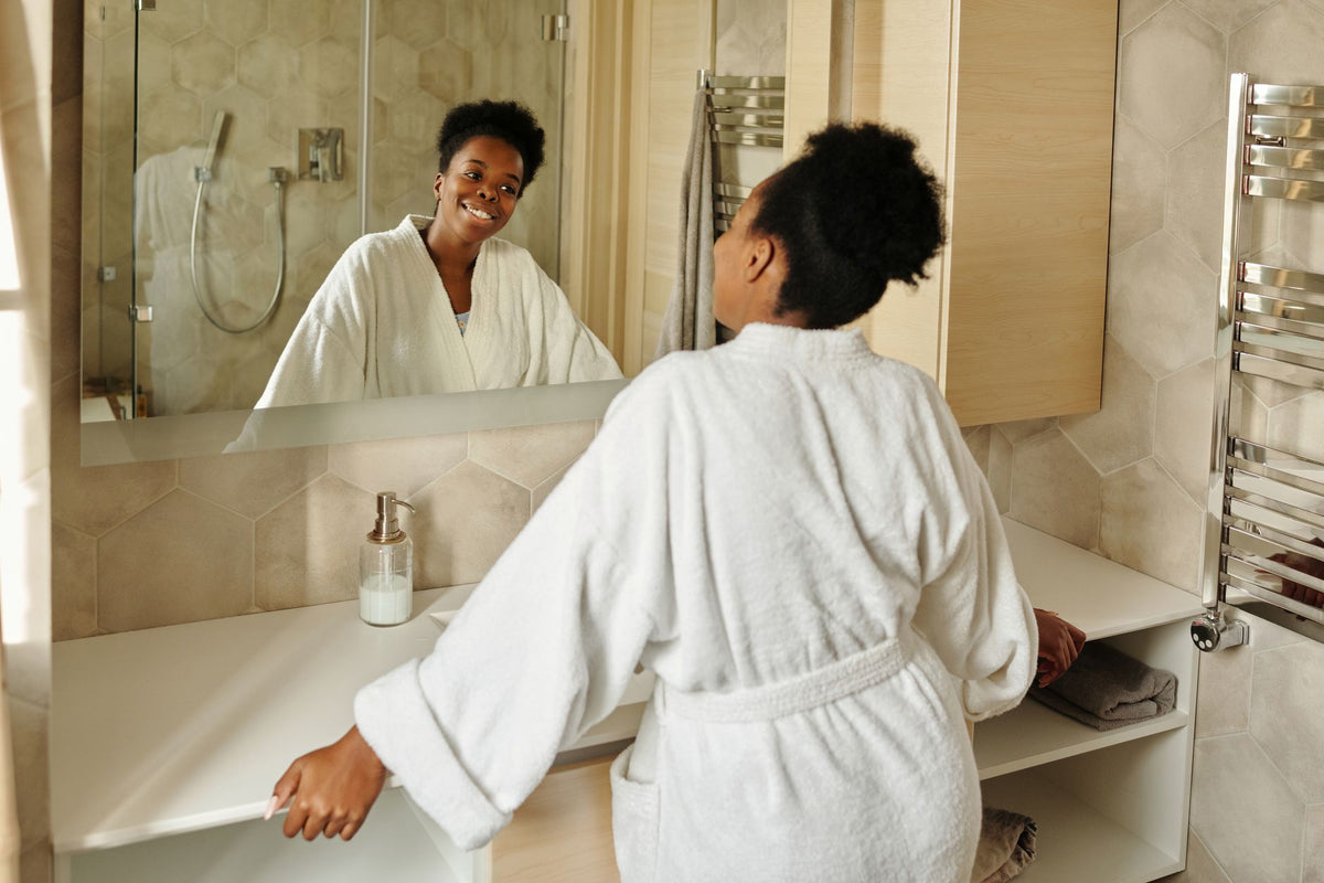 A woman in a bathrobe smiling at her reflection in a modern bathroom mirror