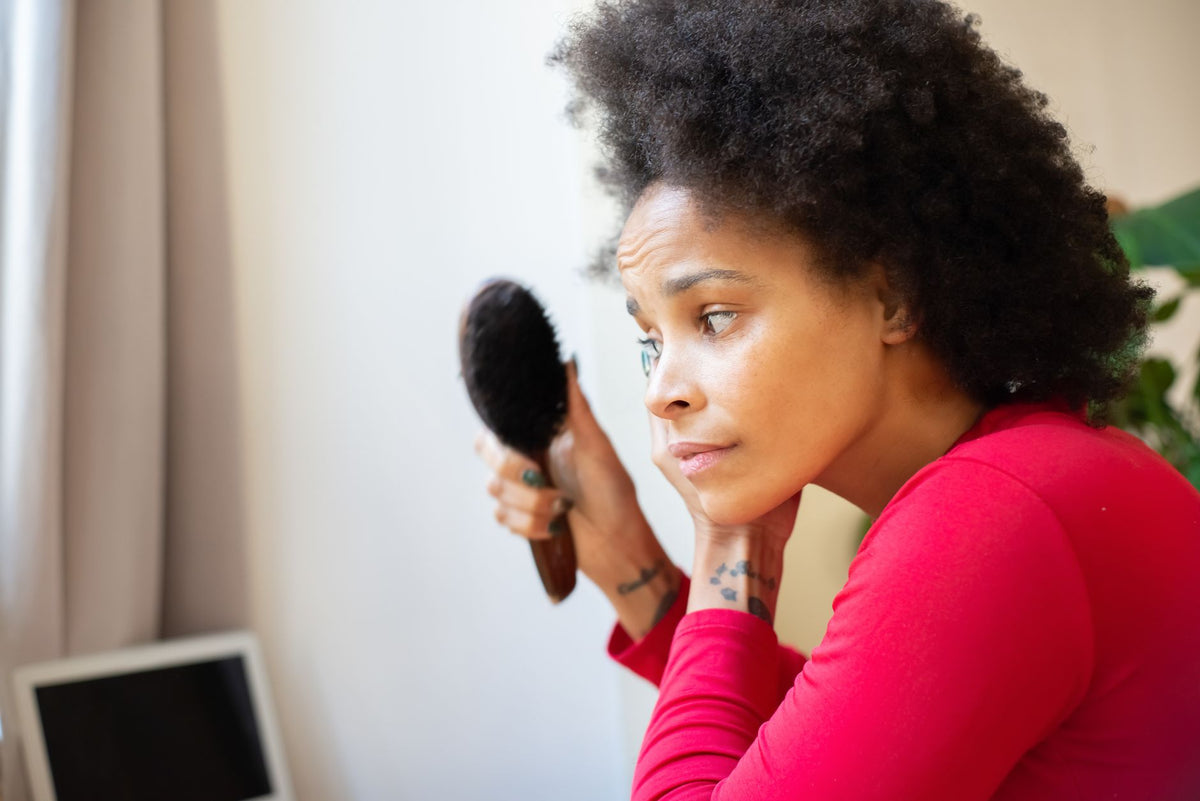 A thoughtful woman with afro hair brushes her hair indoors, reflecting in a personal moment