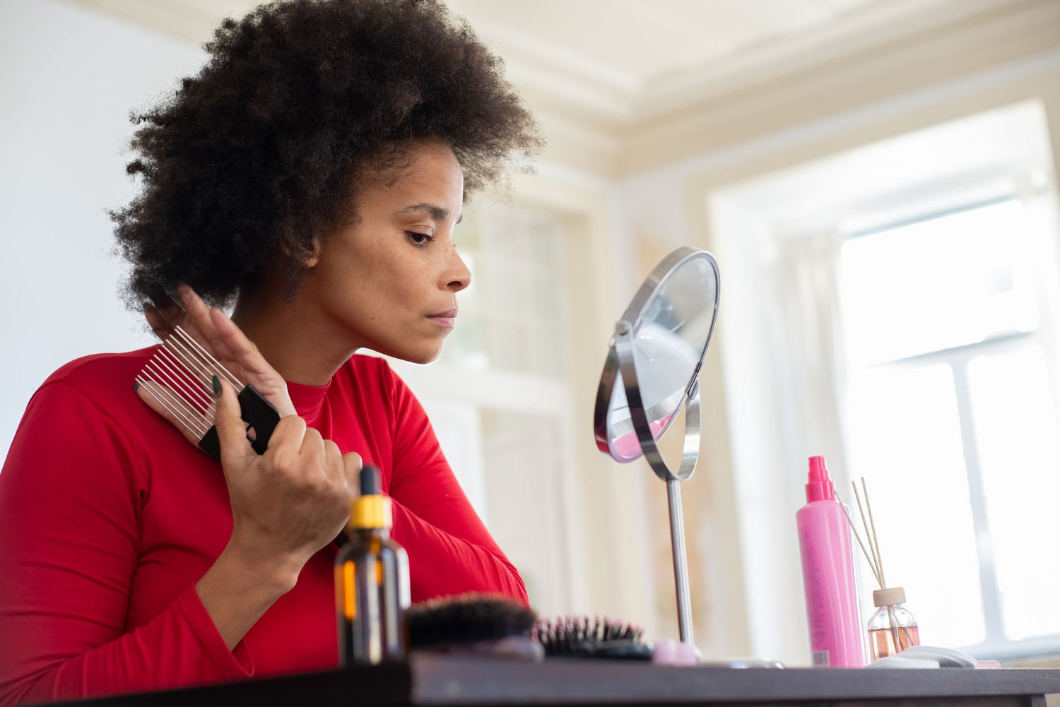 A woman in a red shirt combing her hair by a vanity mirror indoors, creating a personal morning routine scene