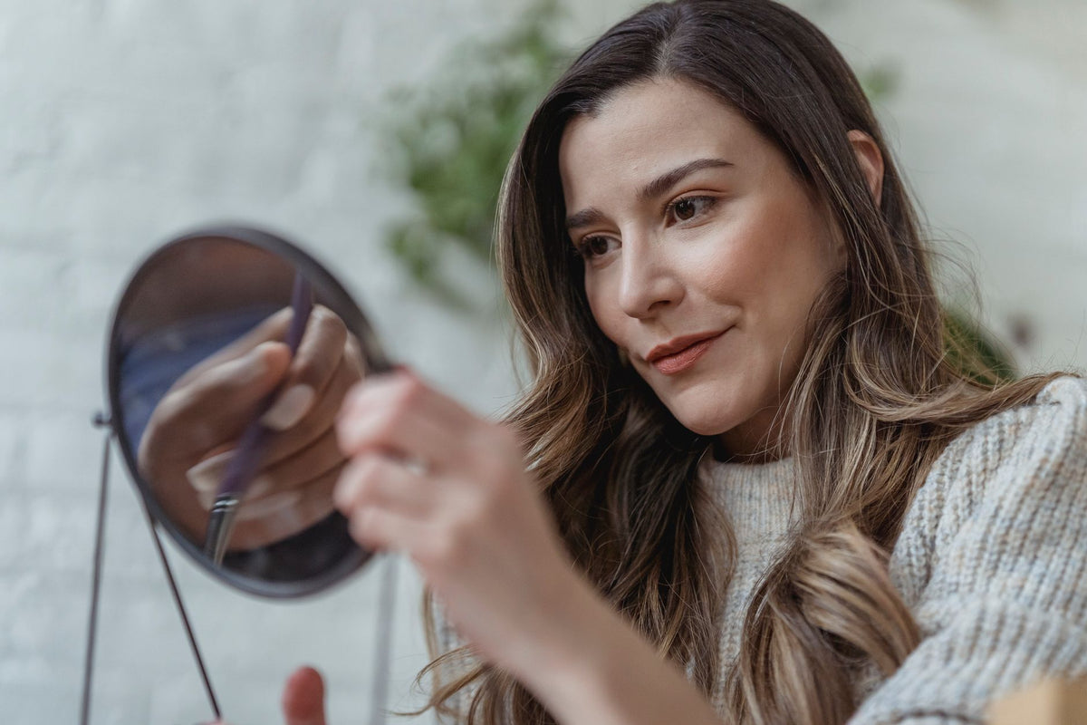 Feminine young woman with long brown wavy hair in stylish sweater looking in round mirror at home in morning