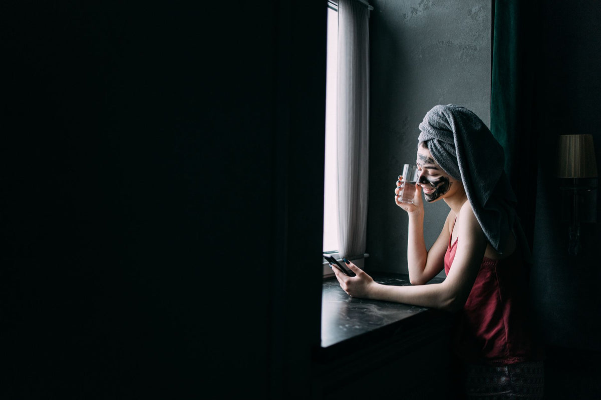 Woman with facial mask and towel turban enjoying skincare routine indoors, holding a glass of water