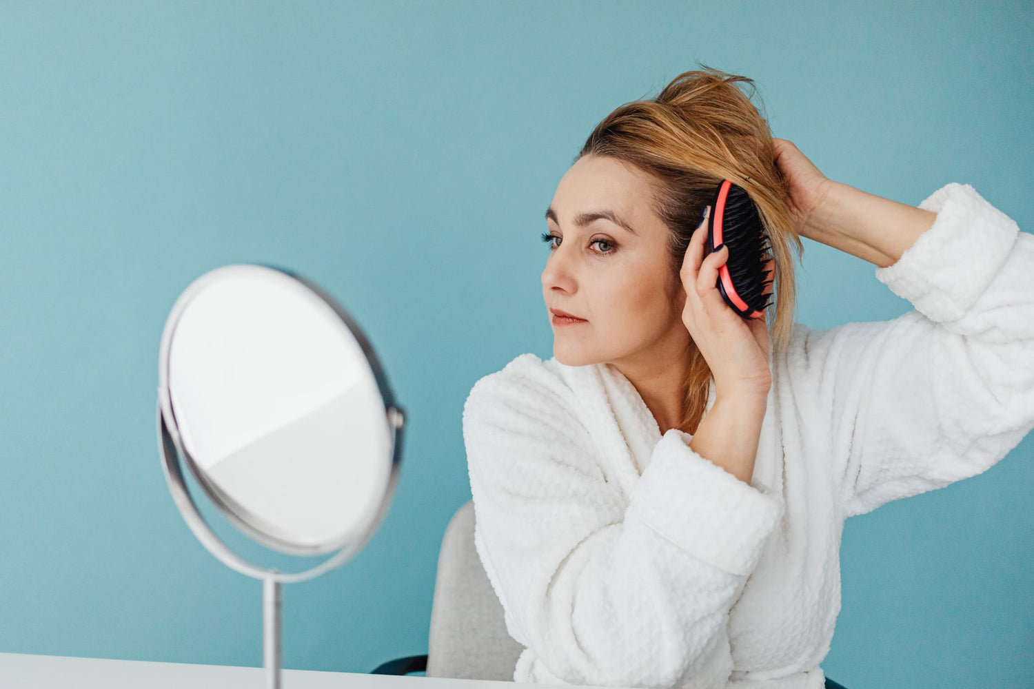 Caucasian woman wearing bathrobe and brushing hair while looking in mirror indoors