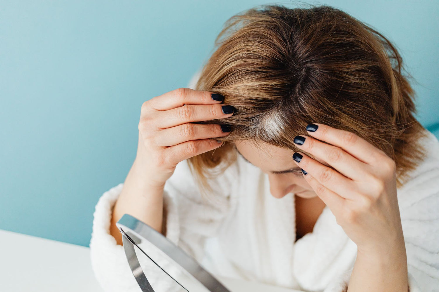 Woman in white robe checking her hair for gray strands using a mirror on blue background