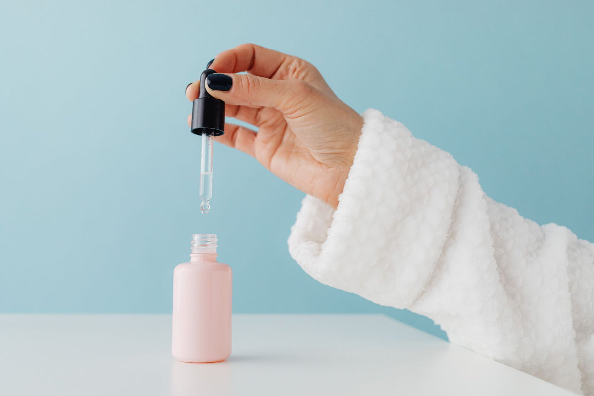 Close-up of a woman's hand in a bathrobe using a serum dropper over a pink glass bottle against a blue background
