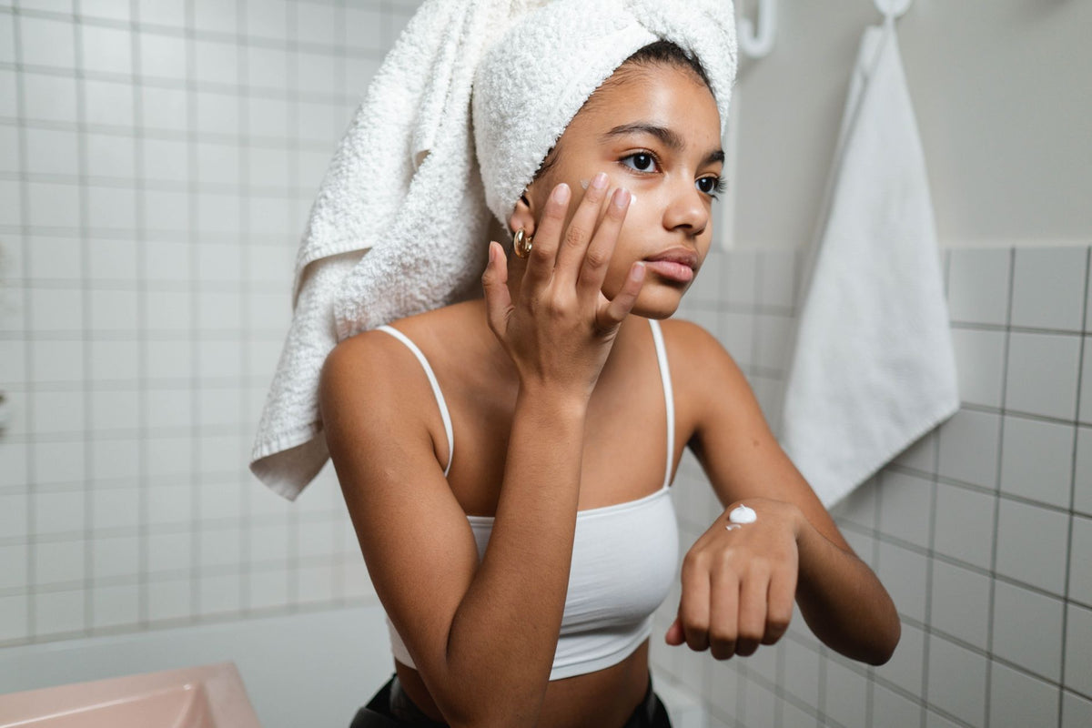 A young woman in a bathroom applying skincare cream with a towel on her head, focusing on self-care