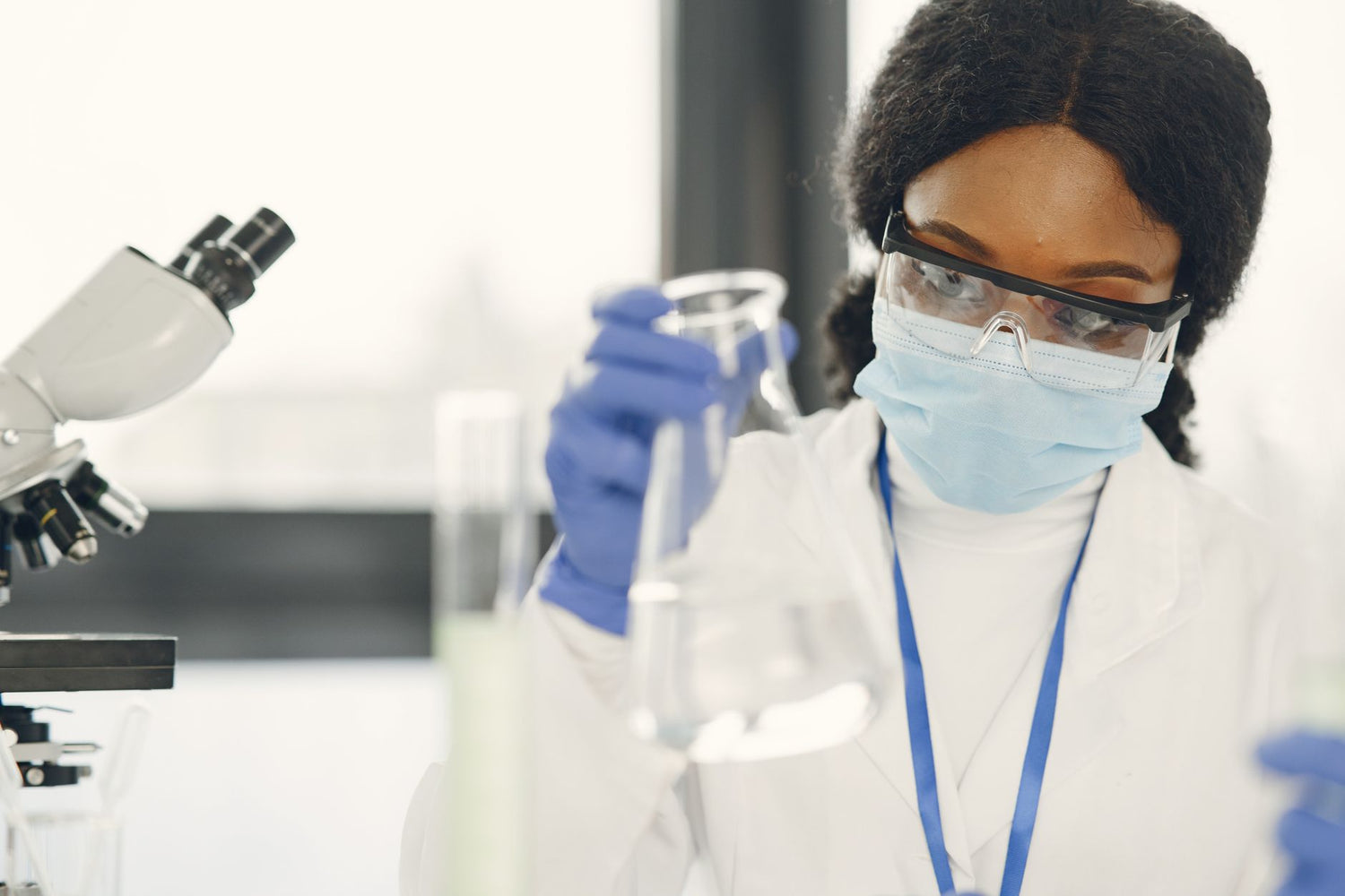 Woman scientist in lab coat and mask conducting experiment with microscope and beaker